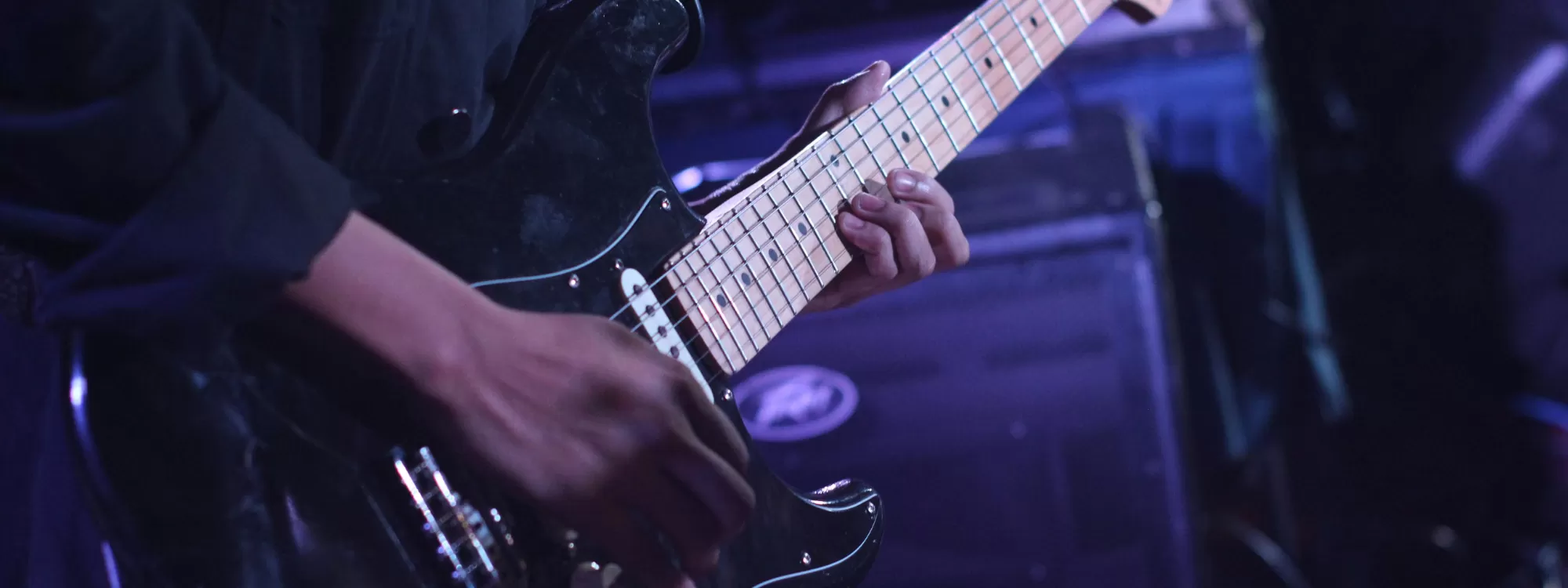 Close-up of a musician playing a black electric guitar during a live performance. The guitarist's hands are visible on the fretboard and strings, illuminated by blue and purple stage lighting